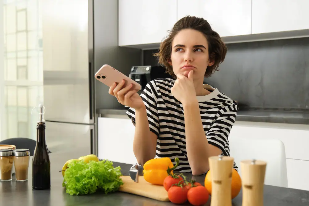 Retrato de uma jovem de pé na cozinha com legumes e uma tábua de cortar com suvida sobre qual dieta seguir